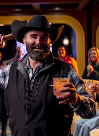 A smiling cowboy pours a drink for himself while posing at a lively bar with friends raising glasses in the background.