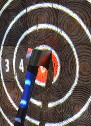 An axe is embedded in the center of a bullseye target on a wooden board marked with concentric circles and numbers.