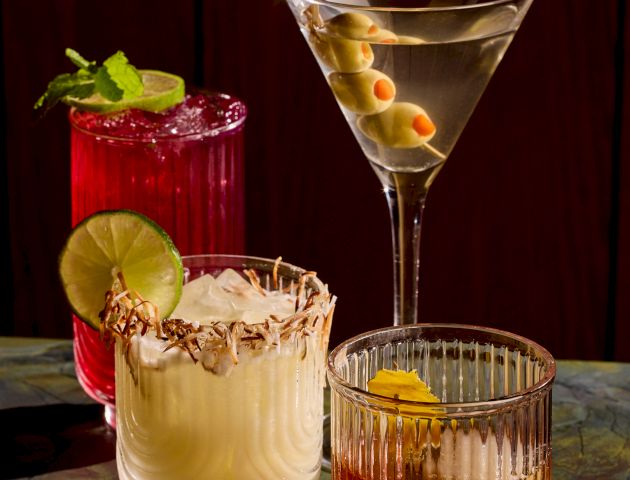 A row of colorful cocktails on a bar, with garnishes like lime and cherry, and elegant glassware in a dim, chic setting.