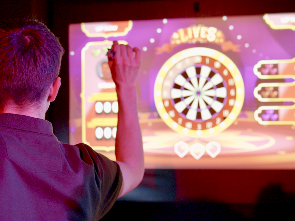 A person is playing a digital darts game on a large screen, showing a dartboard and scoring system.
