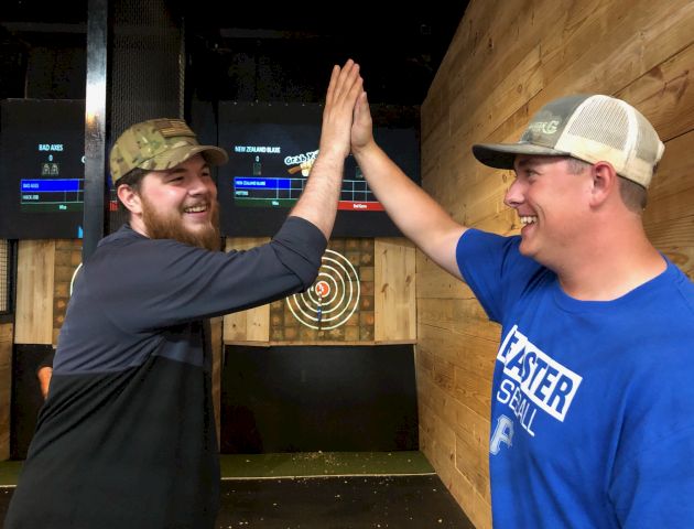 Two people high-fiving in front of a dartboard, smiling, wearing casual hats and clothes, in a recreational setting.