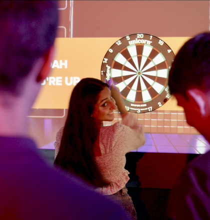 People are playing digital darts in a dimly lit room, with a large dartboard screen displaying scores in the background.