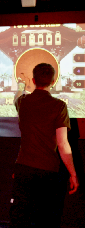 Three people are playing a digital game on a large screen in a dimly lit room with a plant and decor.