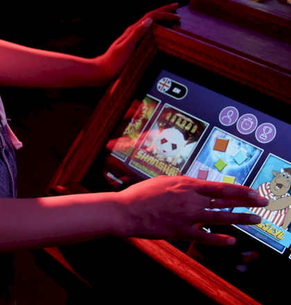A person is using a touchscreen gaming machine, displaying various slot game icons, in a dimly lit setting.