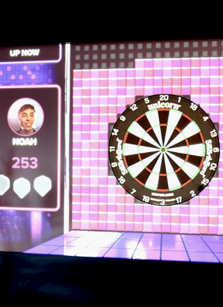The image shows an electronic dartboard game display with player scores and options on a digital screen.