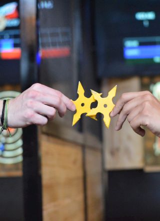 Two hands hold a yellow throwing star with axe-throwing targets in the background, suggesting an activity scene.