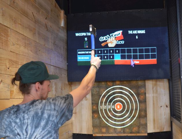 A person is throwing an axe at a target board in an enclosed space; a digital scoreboard displays the results.