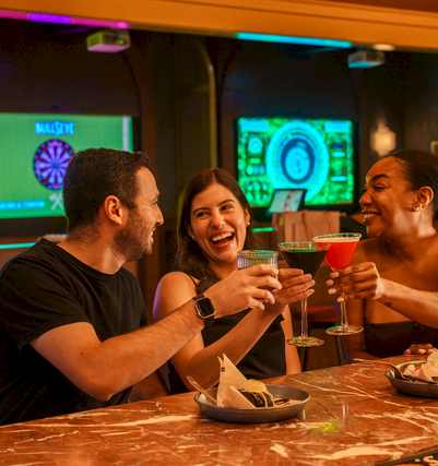 A group of friends at a brightly lit bar cheers with colorful cocktails while gaming screens glow in the background.