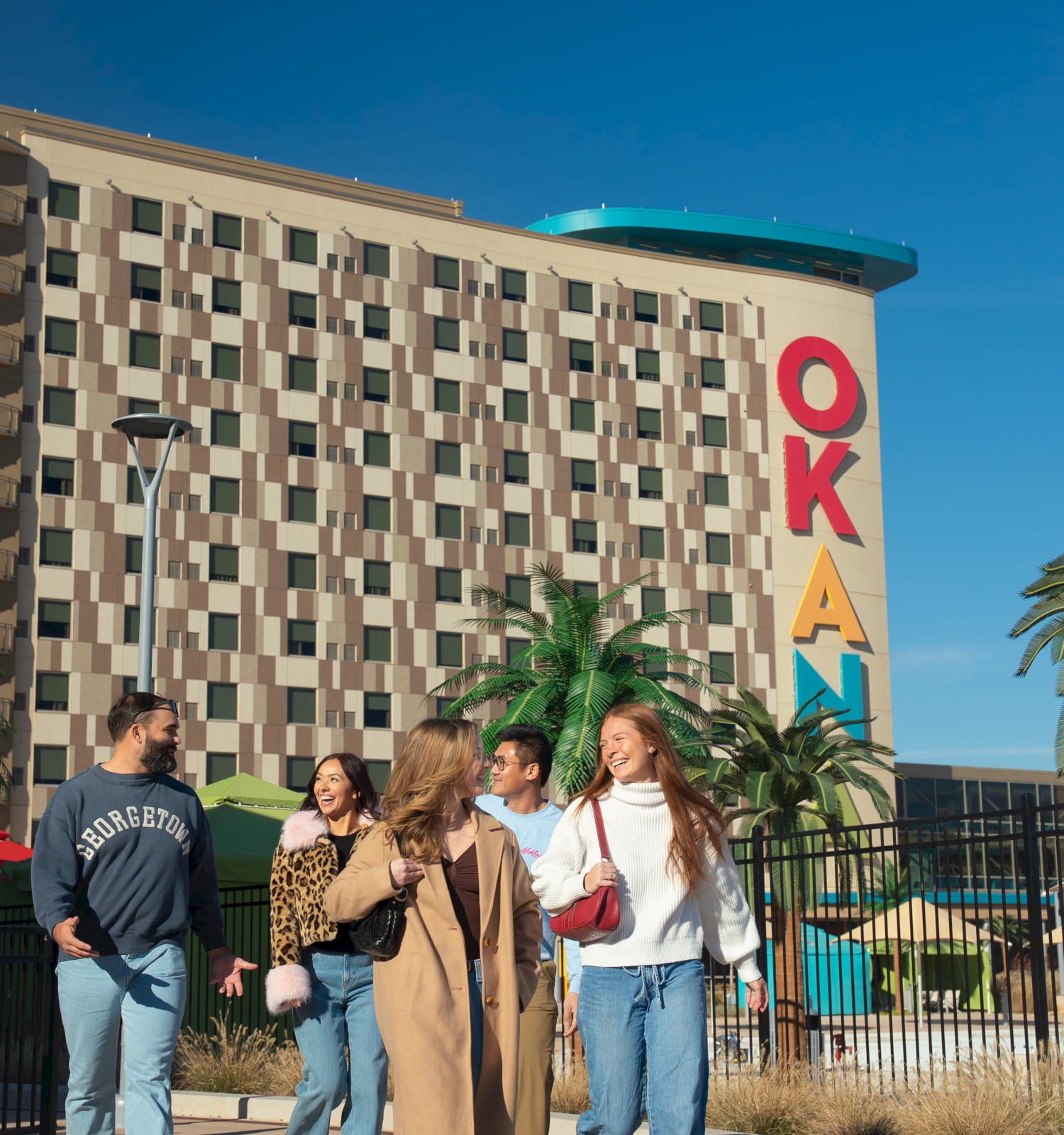 A group of friends walk together outside a colorful hotel named &ldquo;OKA&rdquo; with palm trees and a bright blue sky behind them.
