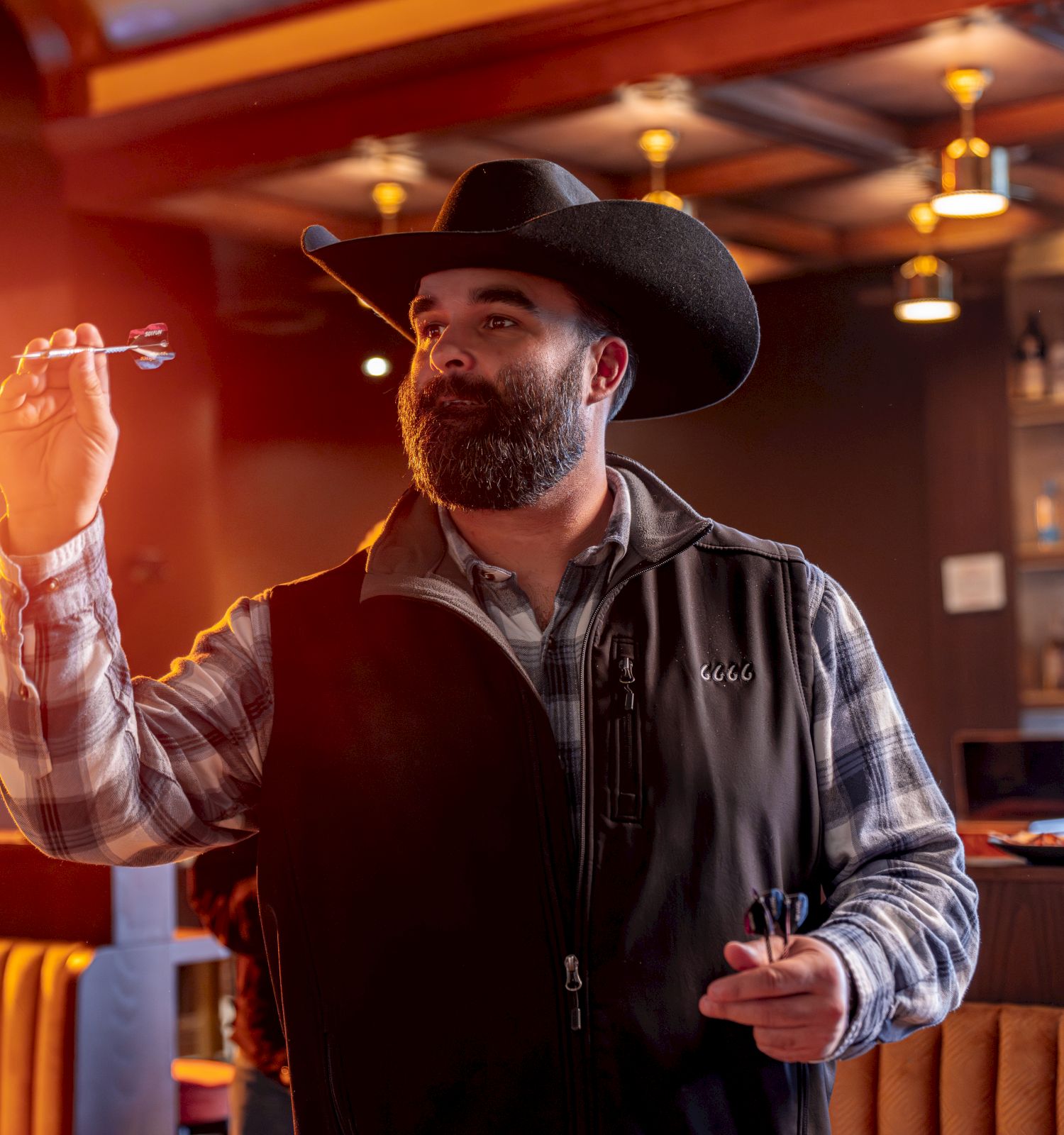 A bearded man in a cowboy hat and vest stands at a bar, gesturing with one hand as he writes or points, warm lights glow around him.
