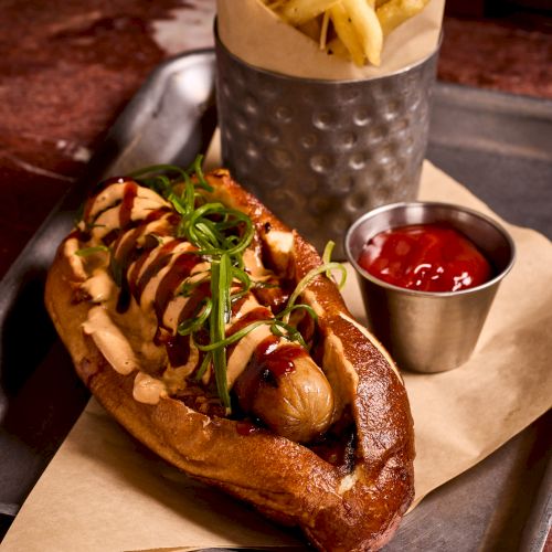 A loaded hot dog with toppings, topped with sauce and herbs, served with fries and a small ketchup cup on the side.