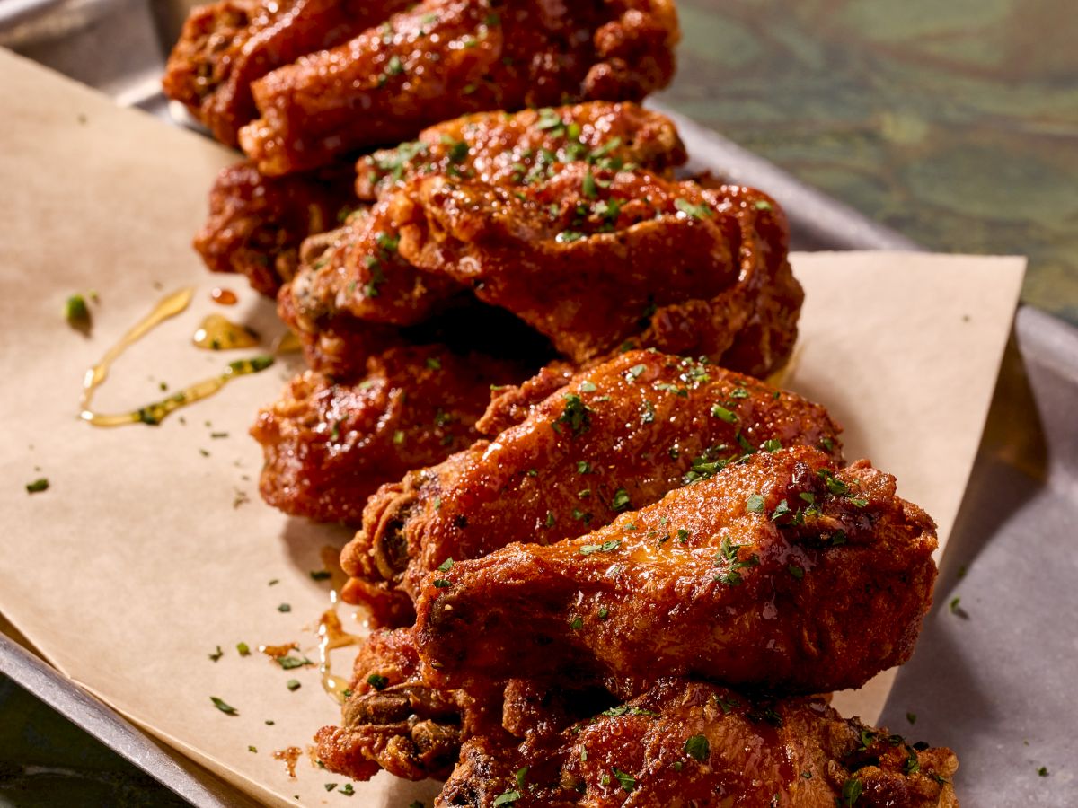 Crispy fried chicken wings on parchment with a dipping sauce, sprinkled with herbs and spices, served in a stack on a metal tray.