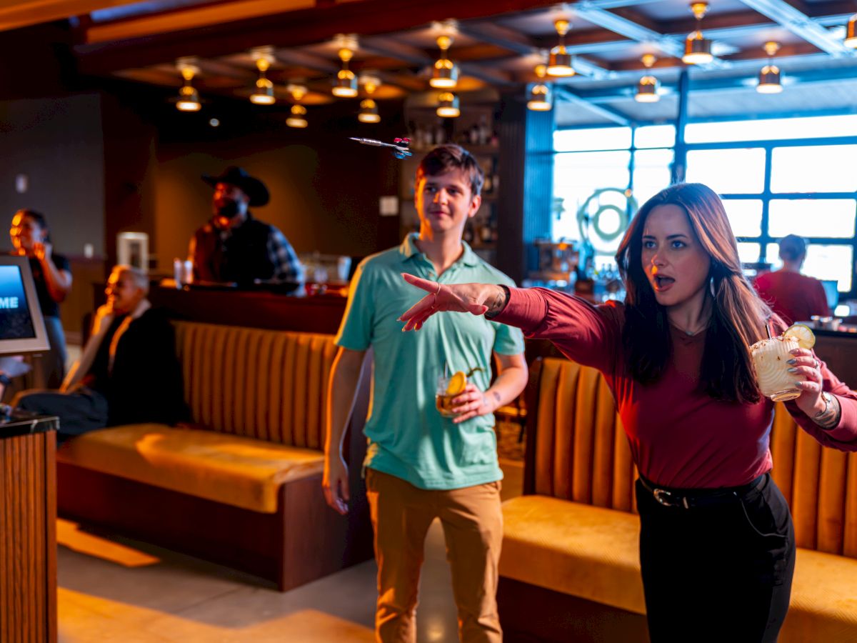 Two people stand in a lively bar or lounge, the woman in a red top gesturing dramatically as the man in a teal shirt watches, with warm lighting.