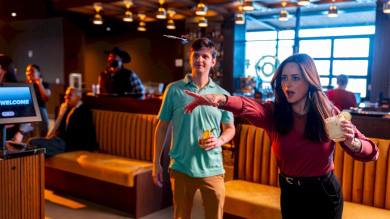 Two people stand in a lively bar or lounge, the woman in a red top gesturing dramatically as the man in a teal shirt watches, with warm lighting.