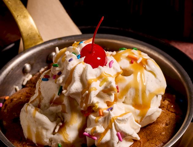 This image looks like a bowl of vanilla ice cream topped with whipped cream and a cherry, possibly with caramel sauce, in a dark bowl.