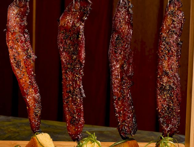 Four strips of dried meat hang on a rod above a wooden board, with small roasted potato chunks garnished with herbs on the board.