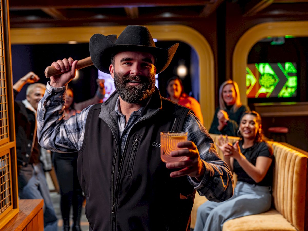 A bearded man in a cowboy hat raises a drink and a pugilistic gesture in a lively bar, with friends cheering in the background.