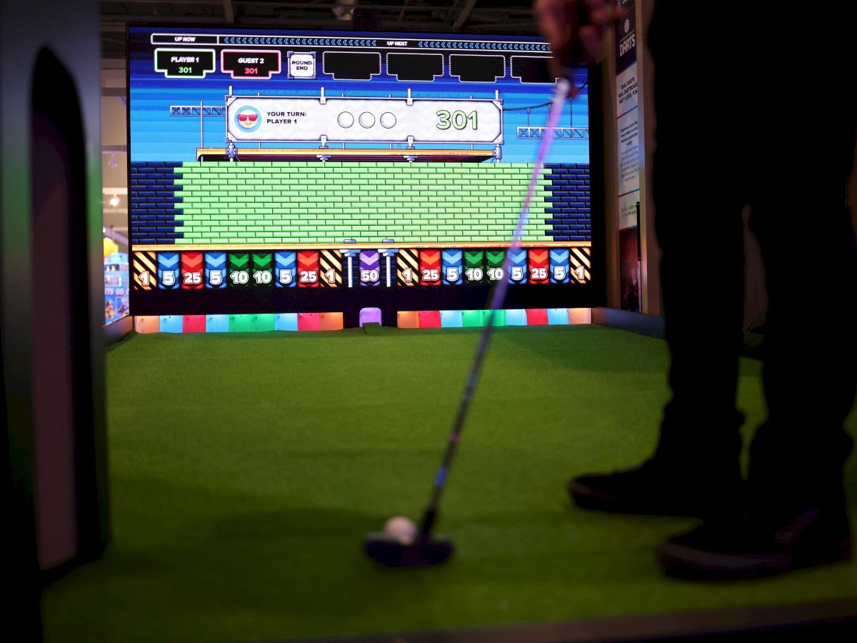 A person playing golf on a virtual mini arcade golf screen at Bullseye in OKC with a bright, retro video game display in the background and a club poised to hit the ball.