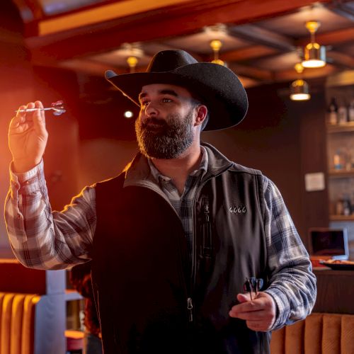 A bearded man in a black cowboy hat and vest gestures with his finger in a warmly lit bar or restaurant.