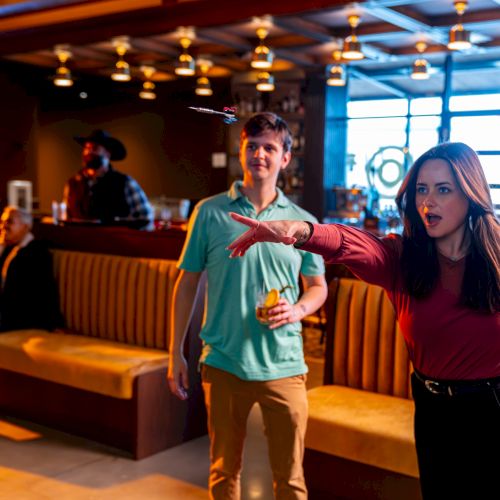 Two people in a lively bowling alley: a woman in red sweater gestures dramatically as a man in teal looks surprised; warm, social vibe.