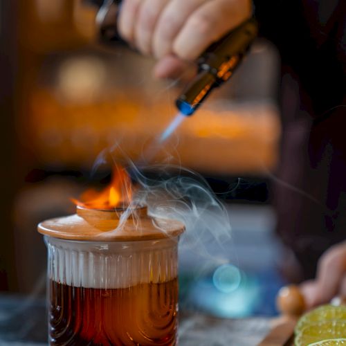 A hand with a torch lights a drink flame atop a glass mug, smoke rising, with lime slices nearby on a counter.