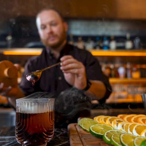 A bartender pouring a dark cocktail; sliced citrus on a board, lime and orange wheels; skewers and glassware on a bar.