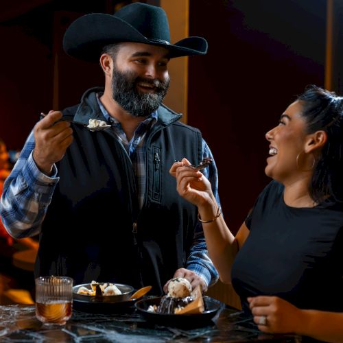 Two people at a bar sharing a laugh, one wearing a cowboy hat and apron, the other smiling while holding a small dessert or bite, good vibes.