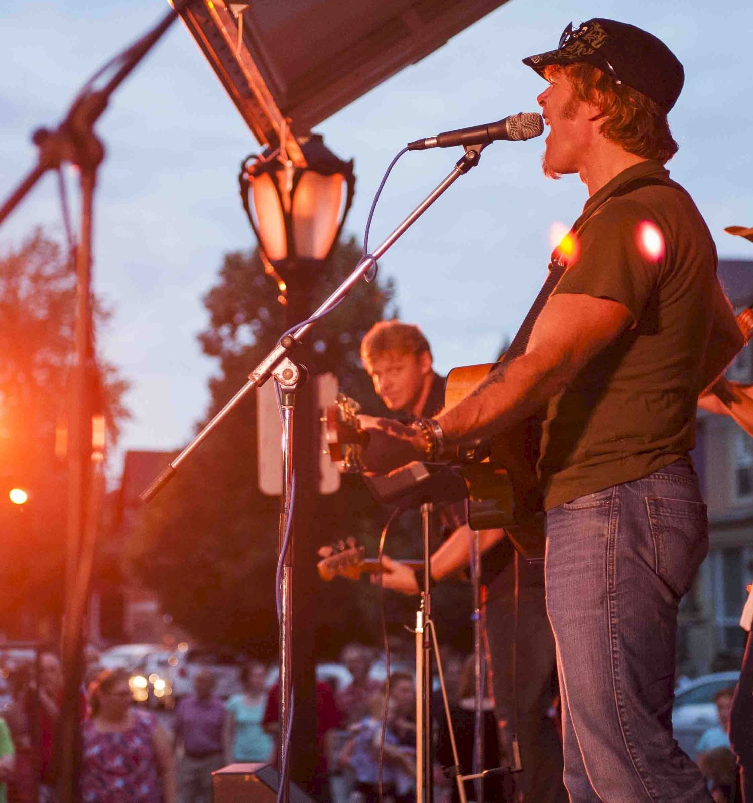 A live band performs on a sunny outdoor stage, guitarist singing into a microphone as fans watch, with warm sunset lights in the background.