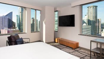 The image shows a modern hotel room with large windows, a bed, a chair, a wall-mounted TV, and a cityscape view outside.