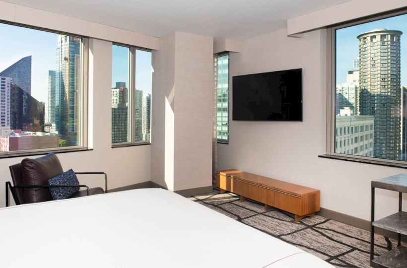 The image shows a modern hotel room with large windows, a bed, a chair, a wall-mounted TV, and a cityscape view outside.