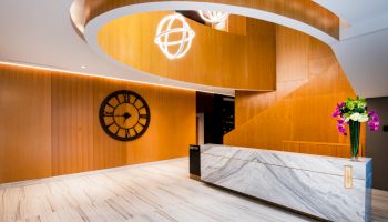 A modern lobby with wooden walls, a large clock, marble reception desk, curved staircase, and decorative lighting and flowers.