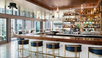 A modern bar with stylish seating and a well-stocked liquor shelf overlooks a cityscape through large windows.