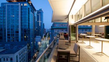 A modern balcony with seating overlooks a cityscape with tall buildings at dusk.