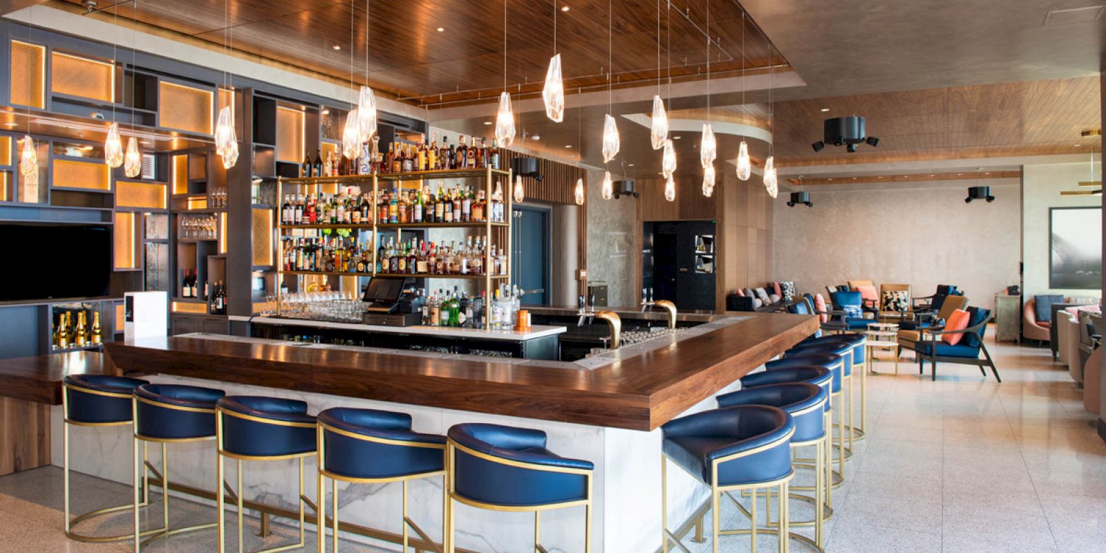 The image shows a modern bar with a wooden ceiling, a countertop, and stylish chairs. Hanging lights create an elegant ambiance.
