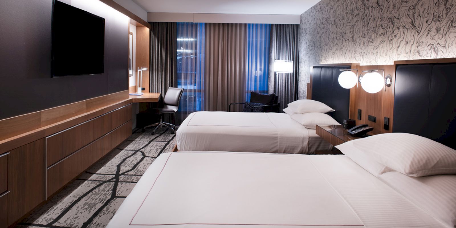 A hotel room with two beds, a wall-mounted TV, a desk, and curtains. The lighting is warm, creating a cozy atmosphere.