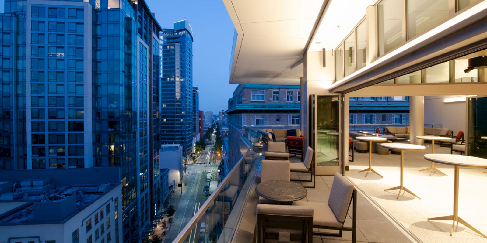 A modern rooftop terrace overlooks a cityscape at dusk, featuring outdoor seating and tables, with nearby high-rise buildings.