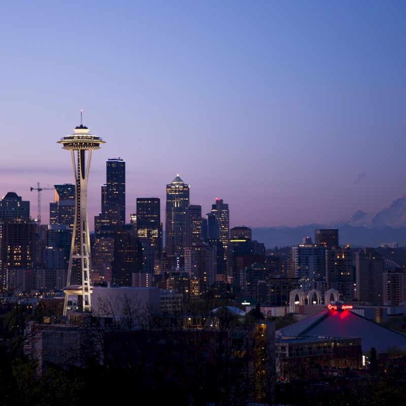 A city skyline at dusk with a prominent tower and mountains in the background, under a slightly cloudy sky.