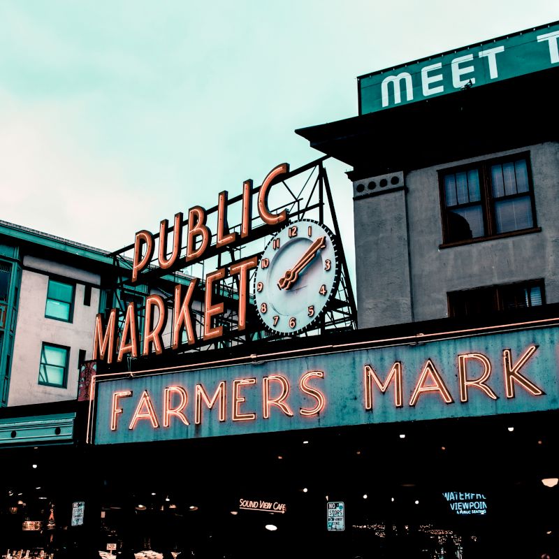 This image shows the iconic sign for the Public Market at Pike Place, featuring a large clock and neon Farmers Market text.