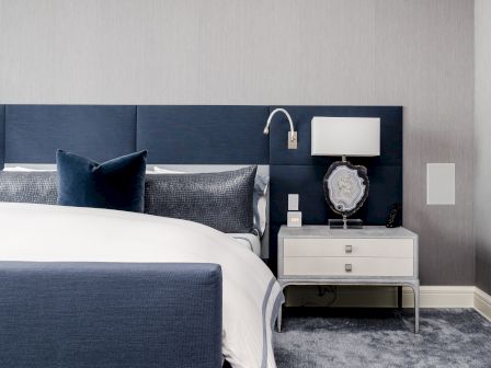 A modern bedroom with a blue headboard, white bedding, gray pillows, and a nightstand with a lamp and decorative items.