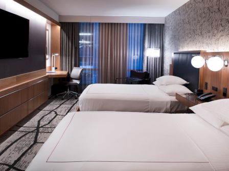 The image shows a hotel room with two beds, a wall-mounted TV, a desk, and lamps by the beds, featuring modern decor.