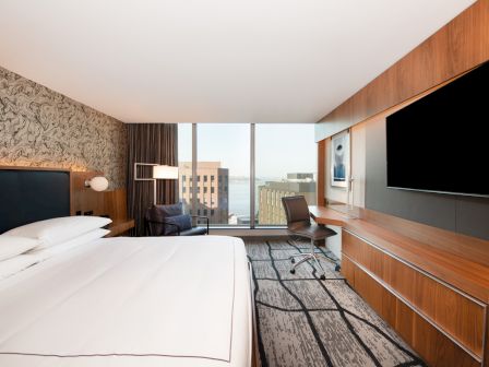 A hotel room with a large bed, wall-mounted TV, desk, chair, and a window overlooking a cityscape.