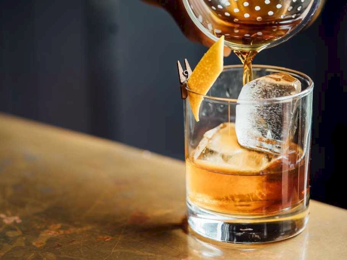 A cocktail is being poured into a glass with ice and a citrus peel garnish on a bar counter.