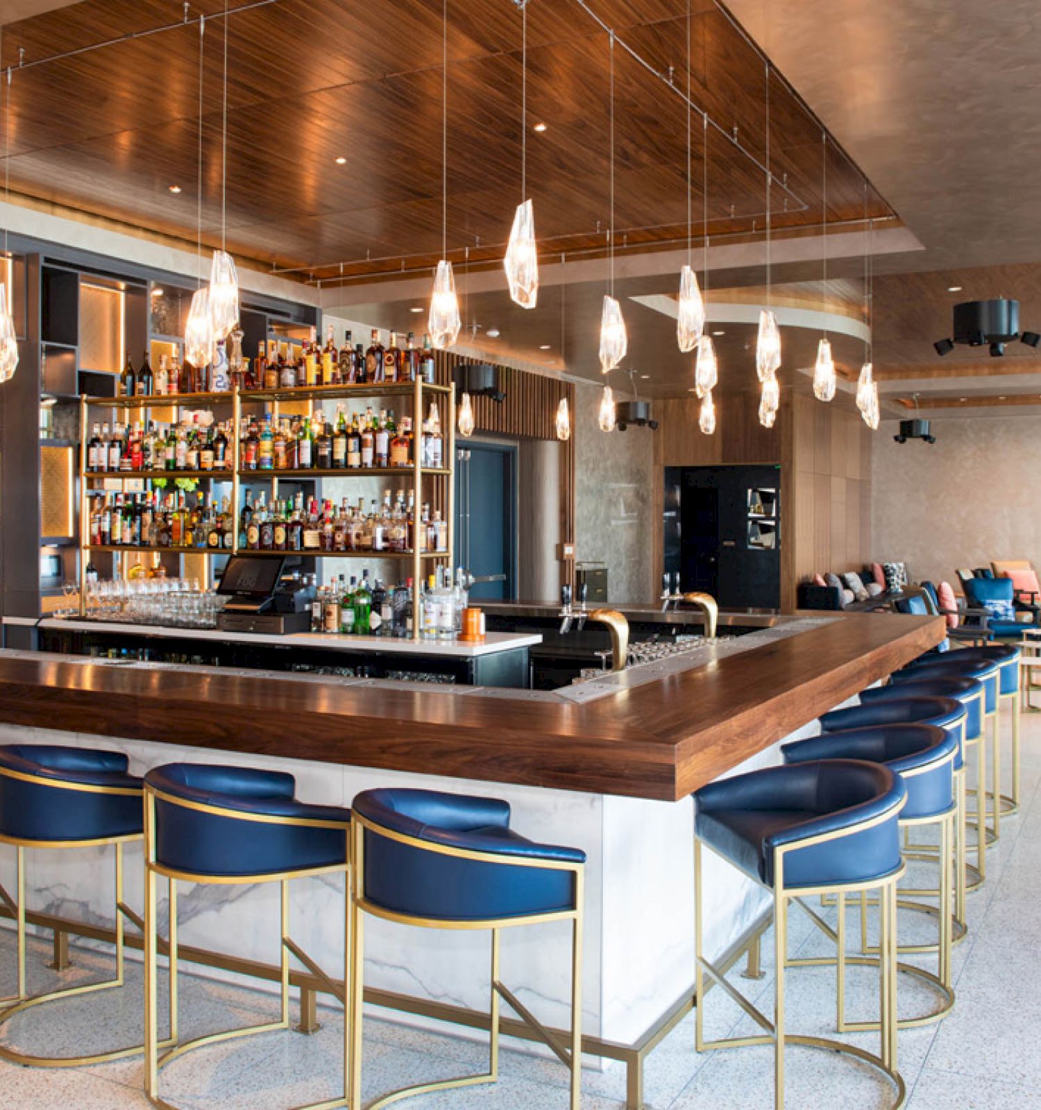The image shows a modern bar with a central counter, blue stools, and pendant lights, surrounded by shelves stocked with bottles.
