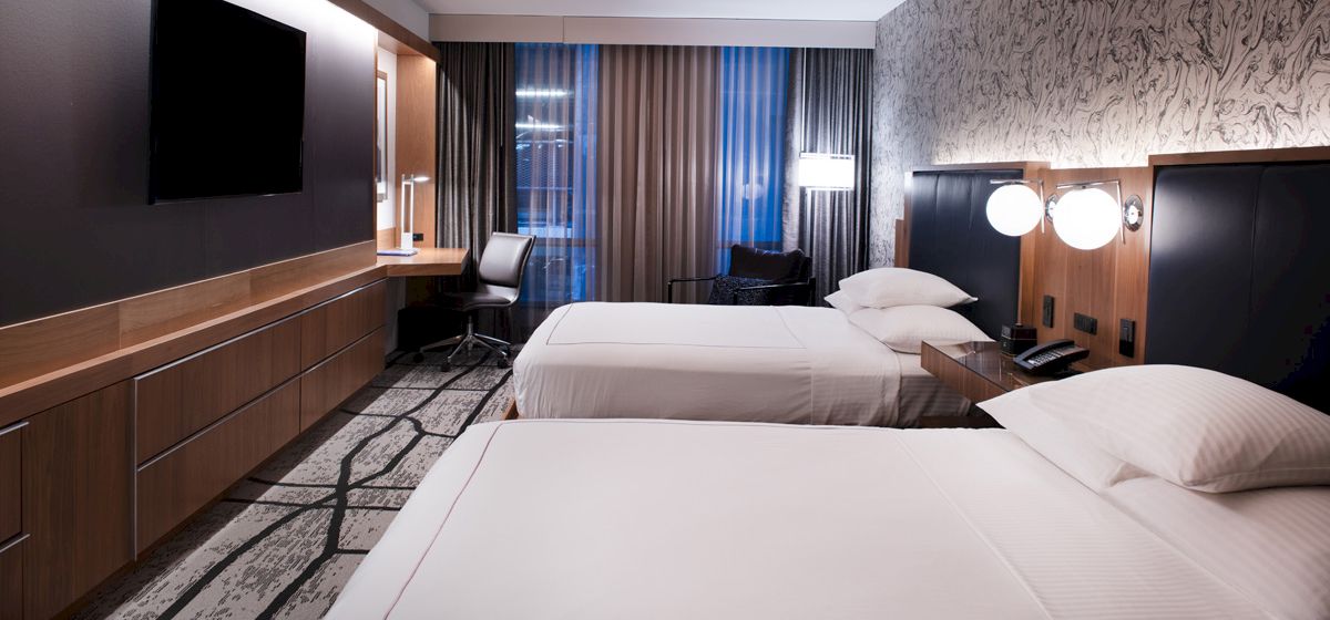 The image shows a modern hotel room with two beds, a TV, a desk, and lamps, featuring a stylish design and ample natural light.