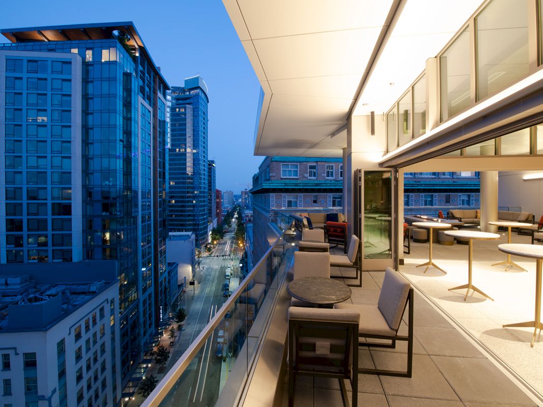 A modern rooftop terrace overlooks a cityscape with tall buildings, featuring seating areas and tables under ambient lighting.