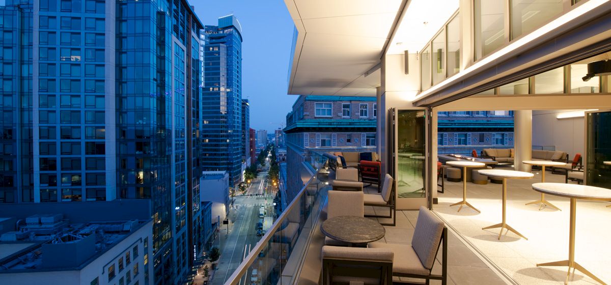 A modern rooftop terrace overlooks a cityscape with tall buildings, featuring seating areas and tables under ambient lighting.