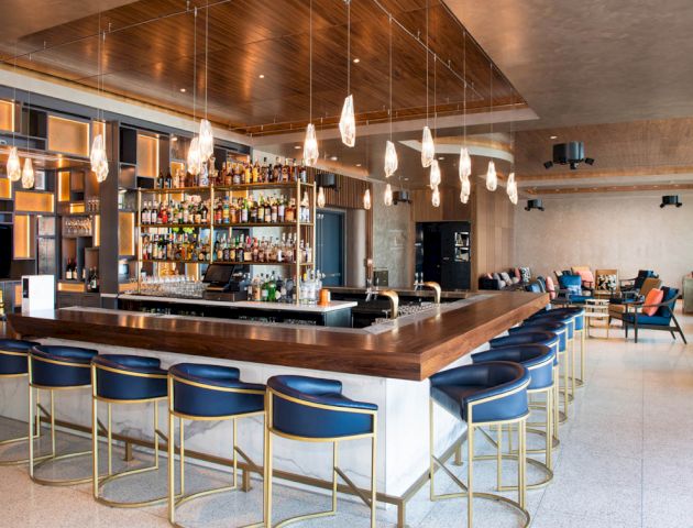 The image shows a modern bar interior with blue stools, wooden accents, pendant lights, and a fully stocked bar area.