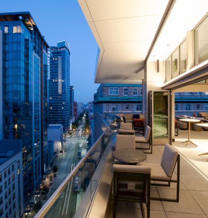 A modern cityscape at twilight showing a high-rise balcony with seating and tables overlooking a street lined with buildings.