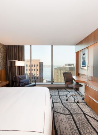A modern hotel room with a large bed, wall-mounted TV, desk, and window with a city view.