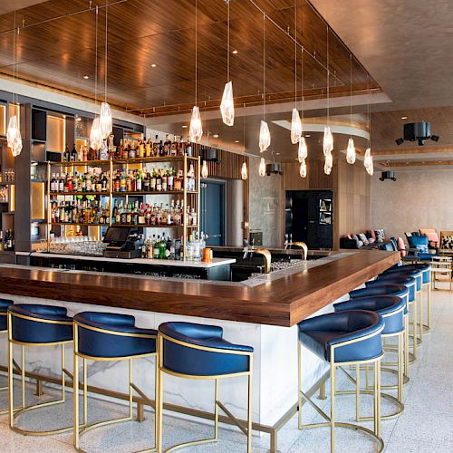 The image shows a modern bar with suspended lights, a stocked liquor shelf, and blue cushioned stools around the counter.
